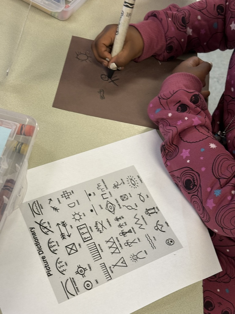 A close-up of a student’s hands drawing Native American symbols on brown paper, using a “Picture Dictionary” guide sheet with pictographs for reference.