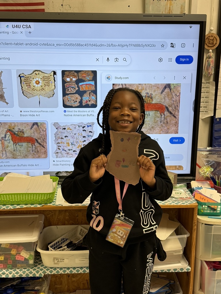 A smiling student holds up a brown paper “buffalo hide” drawing featuring small symbols and figures, created as part of a Native American heritage lesson.