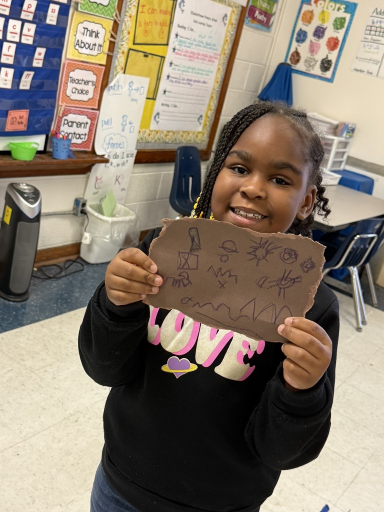 A student smiles while holding their completed buffalo hide artwork decorated with symbols and scenes, standing in a classroom with colorful posters on the wall.