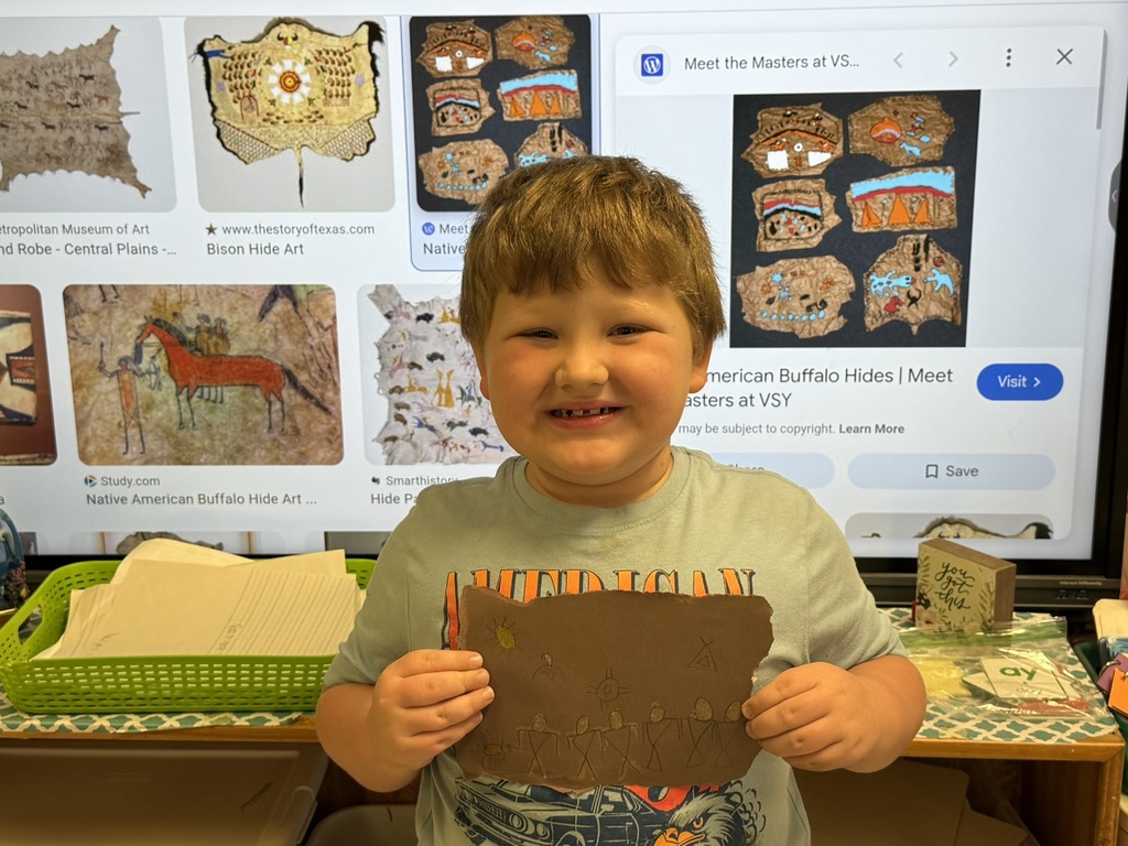 A smiling student holds a brown paper “buffalo hide” artwork decorated with Native American-style symbols, with images of authentic buffalo hide paintings displayed on the screen behind him.