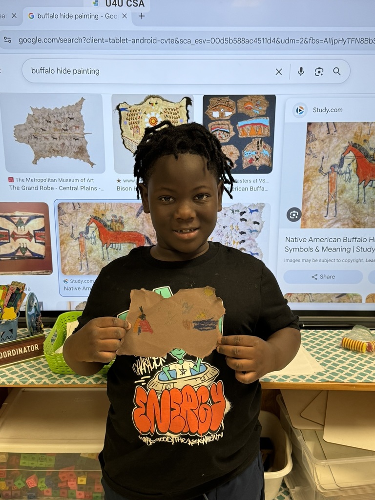 A student proudly shows their completed “buffalo hide painting” project with colorful drawings inspired by Native American art, standing in front of a classroom screen showing buffalo hide art examples.
