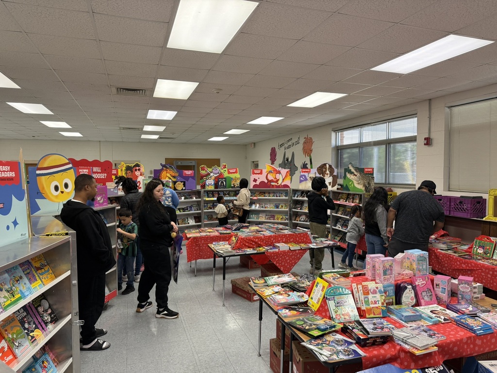 School library set up for the Scholastic Book Fair with red-covered tables and bookcases full of books and gift items. Families and students browse the displays around the room.