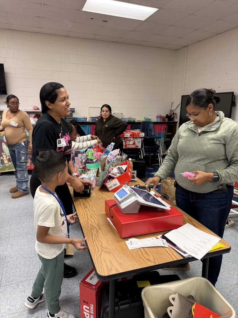 A staff member helps a parent and student at the book fair checkout table with a card reader, baskets of small items, and receipts.