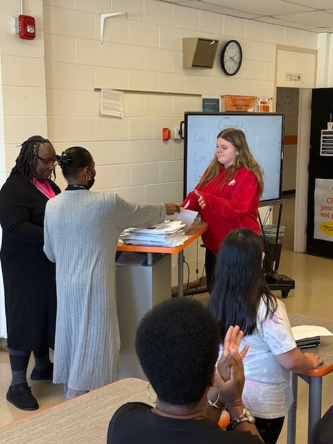 A teacher stands at the front of a classroom handing out certificates to students while others applaud. A digital screen and classroom materials are visible in the background.