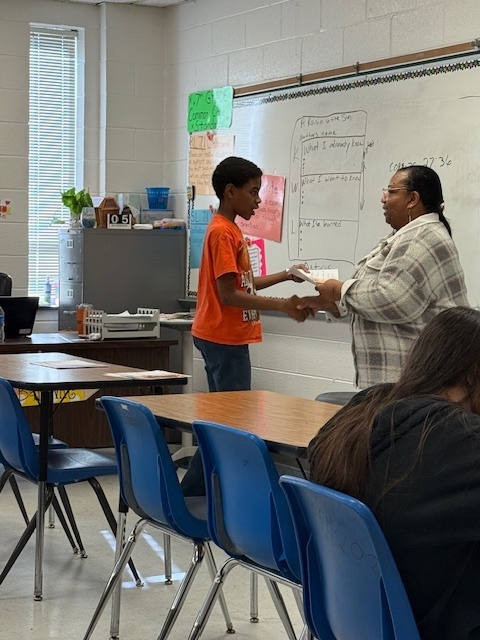 A student in an orange shirt receives an award certificate from a teacher near the whiteboard, with classmates watching from their desks.