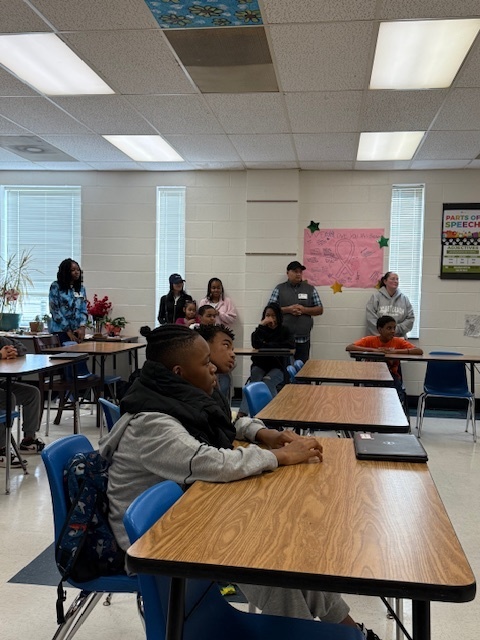 Students and family members sit attentively in a classroom decorated with posters and flowers during the awards ceremony.