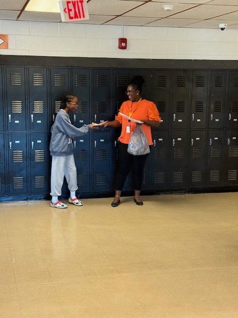 A teacher stands by the hallway lockers presenting an award to a student dressed in gray, both smiling during the recognition.