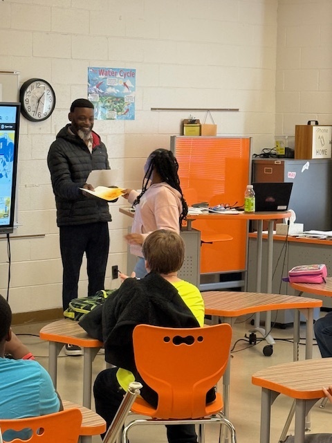 A teacher and student stand at the front of a classroom while the student receives a certificate; other students are seated at their desks watching.