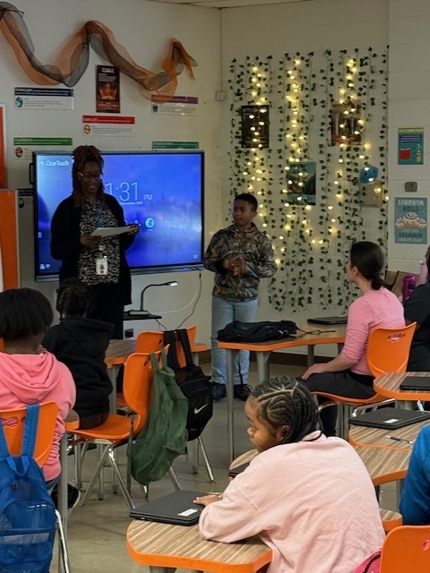 A teacher presents an award to a student in a classroom decorated with string lights and green vines while classmates watch from their desks.