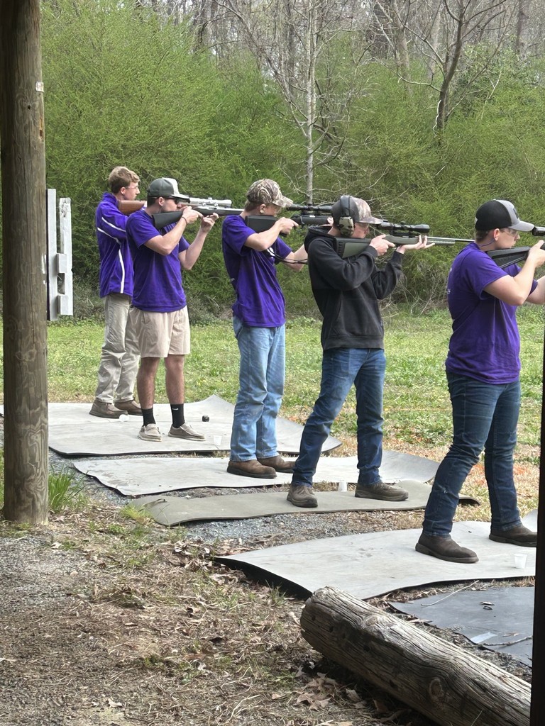 Five students in purple shirts practice rifle shooting outdoors at a range surrounded by trees.