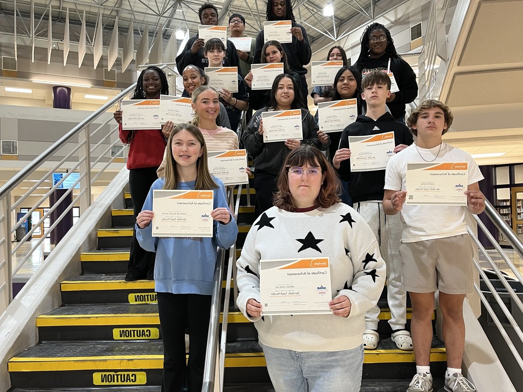 Students stand on school stairs holding certificates of achievement, smiling and posing for a group photo indoors.
