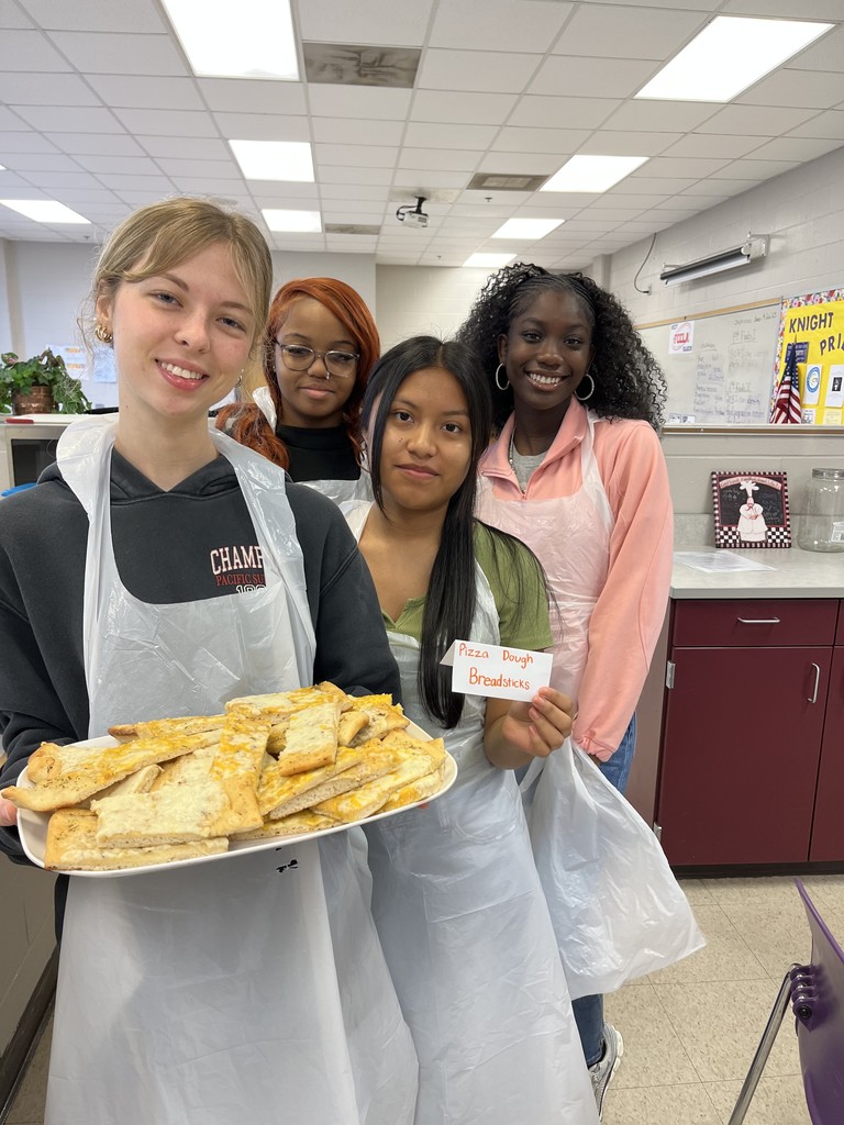 Four smiling students in plastic aprons holding a platter of breadsticks and a sign reading "Pizza Dough Breadsticks.