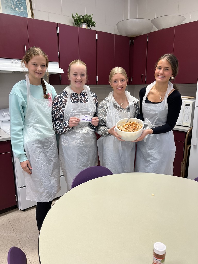 Four smiling students in plastic aprons in a kitchen, holding a large bowl of corn salad and a sign reading "Corn Salad.