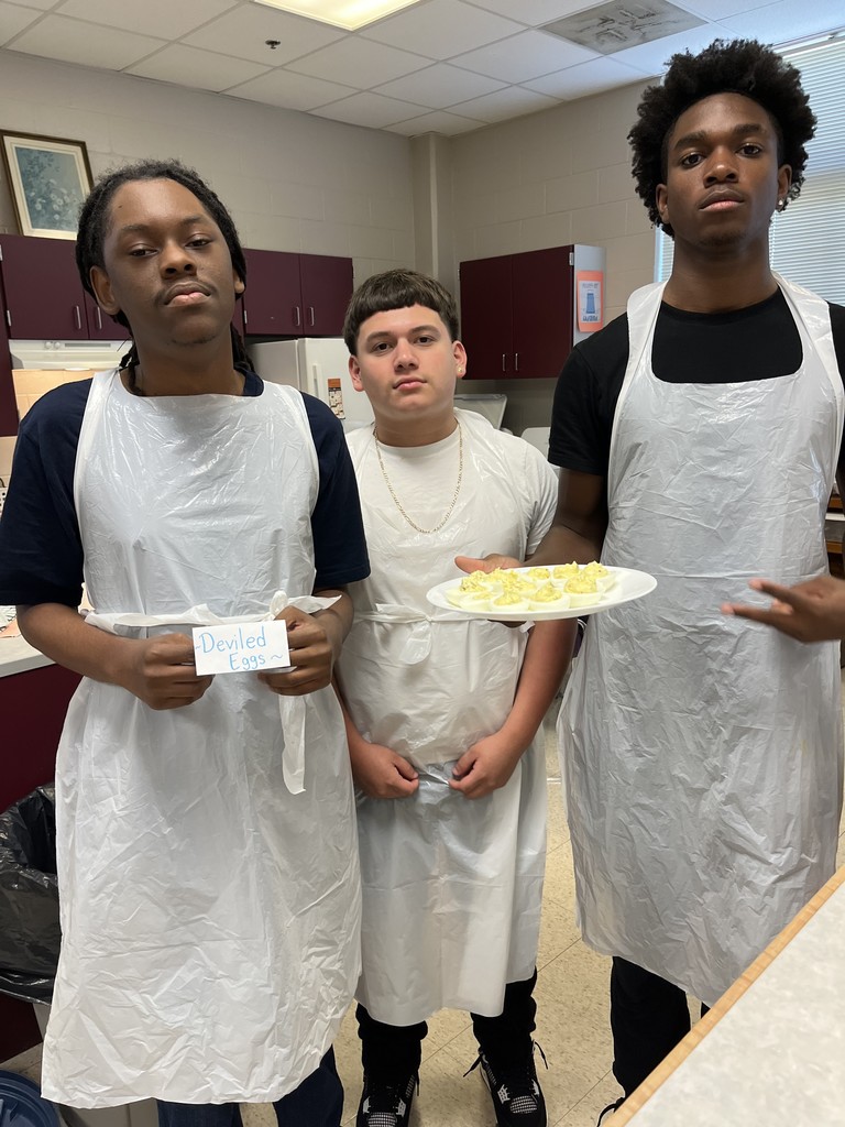 Three students in plastic aprons holding a plate of deviled eggs and a sign reading "Deviled Eggs.