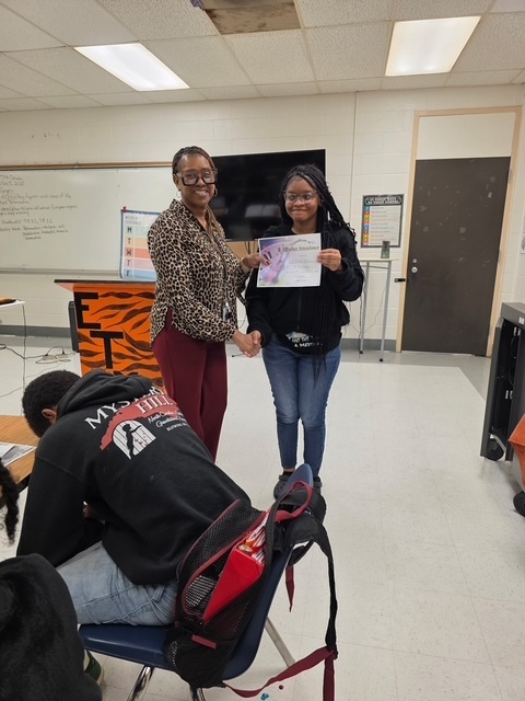 A teacher shakes hands with a student who is holding a certificate. Both are smiling as they celebrate the student’s achievement.