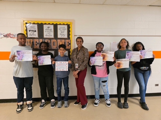 A group of students stand in a hallway with their teacher, each holding certificates for their first quarter awards. A “Welcome” bulletin board and motivational posters decorate the wall behind them.