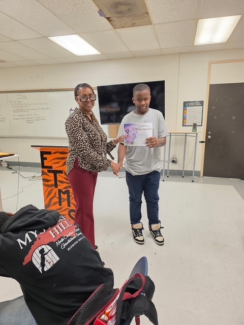 A teacher and student stand side by side in the classroom, smiling and holding an award certificate during the first quarter recognition ceremony.