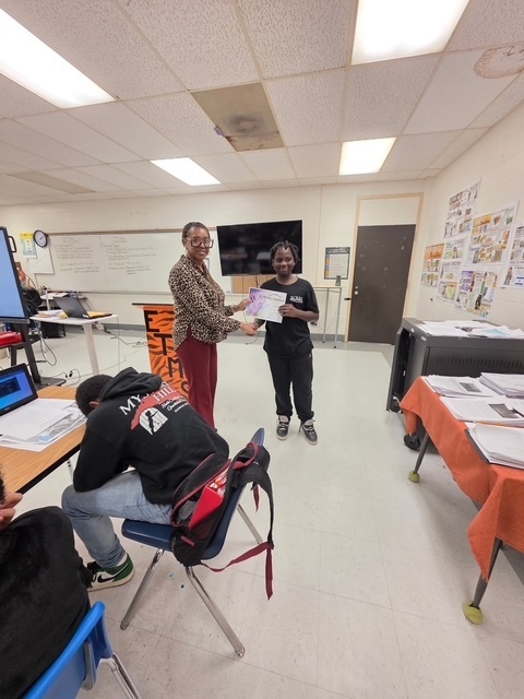 A teacher smiles as she presents a certificate to a student standing at the front of the classroom. The student proudly holds up their award while classmates look on.