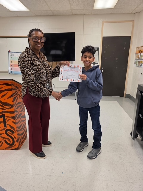 A student in a gray hoodie stands beside a teacher, holding a “Student of the Month” certificate and smiling for the camera.