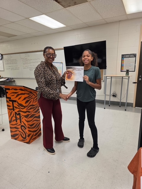 A student in a green shirt smiles while receiving an award certificate from a teacher at the front of the classroom.