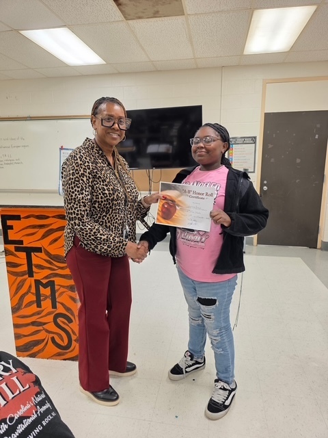 A teacher presents an A/B Honor Roll certificate to a smiling student dressed in pink, standing proudly in front of the classroom.