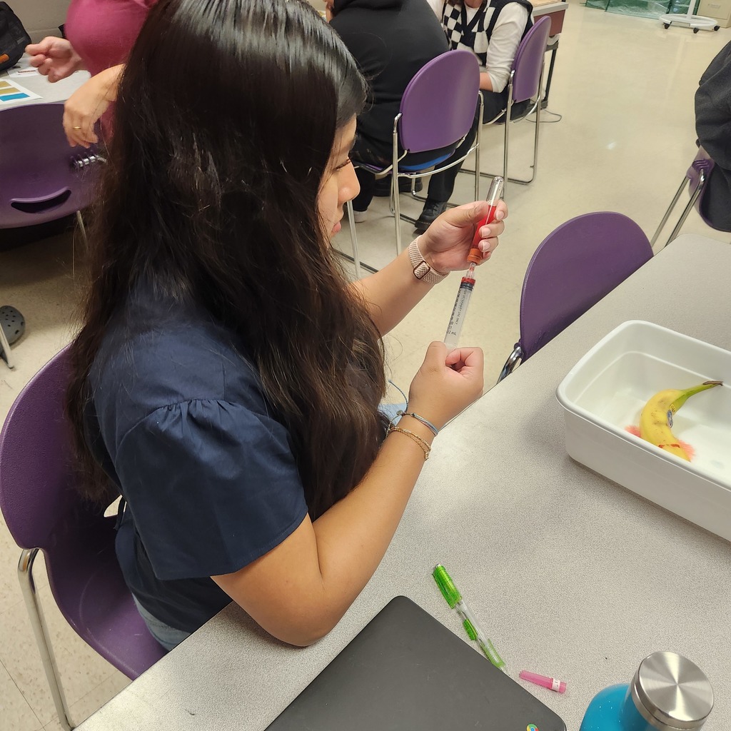 A student holds a large syringe with red liquid, working on a classroom activity with a banana in a tray.