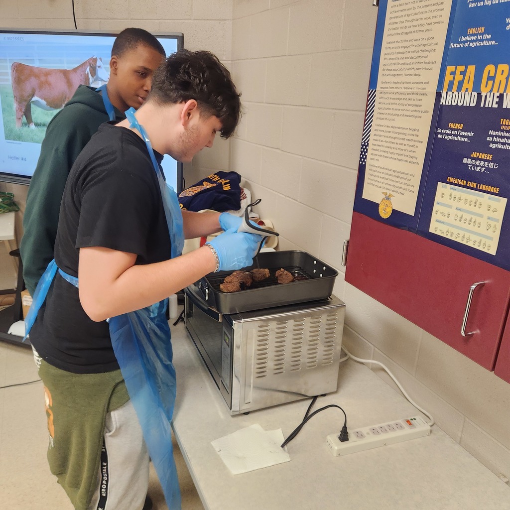Two students, wearing aprons, grill food on a counter. A screen shows an image of cattle behind them