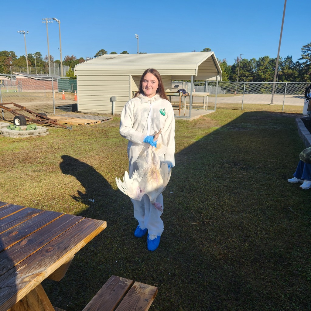 A smiling student in a white protective suit and blue gloves holds a poultry bird outside near a shed