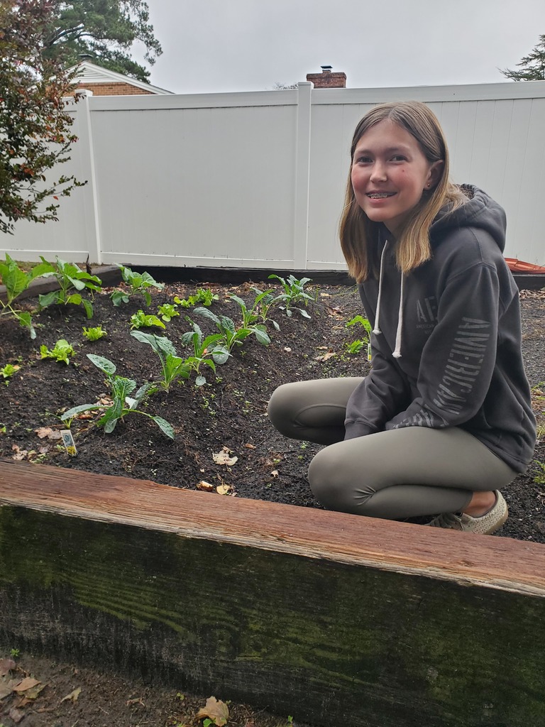 A smiling young person in a dark hoodie and green leggings kneels beside a raised garden bed filled with small plants.