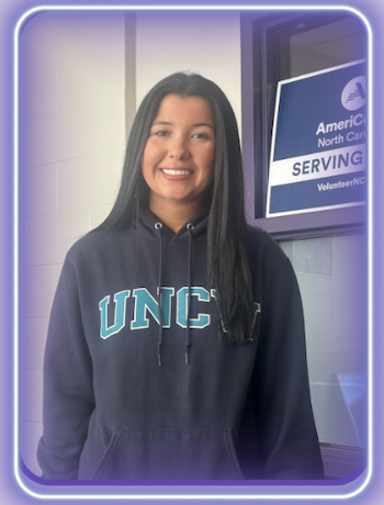 A smiling young woman in a UNCW hoodie stands in front of an AmeriCorps North Carolina sign.