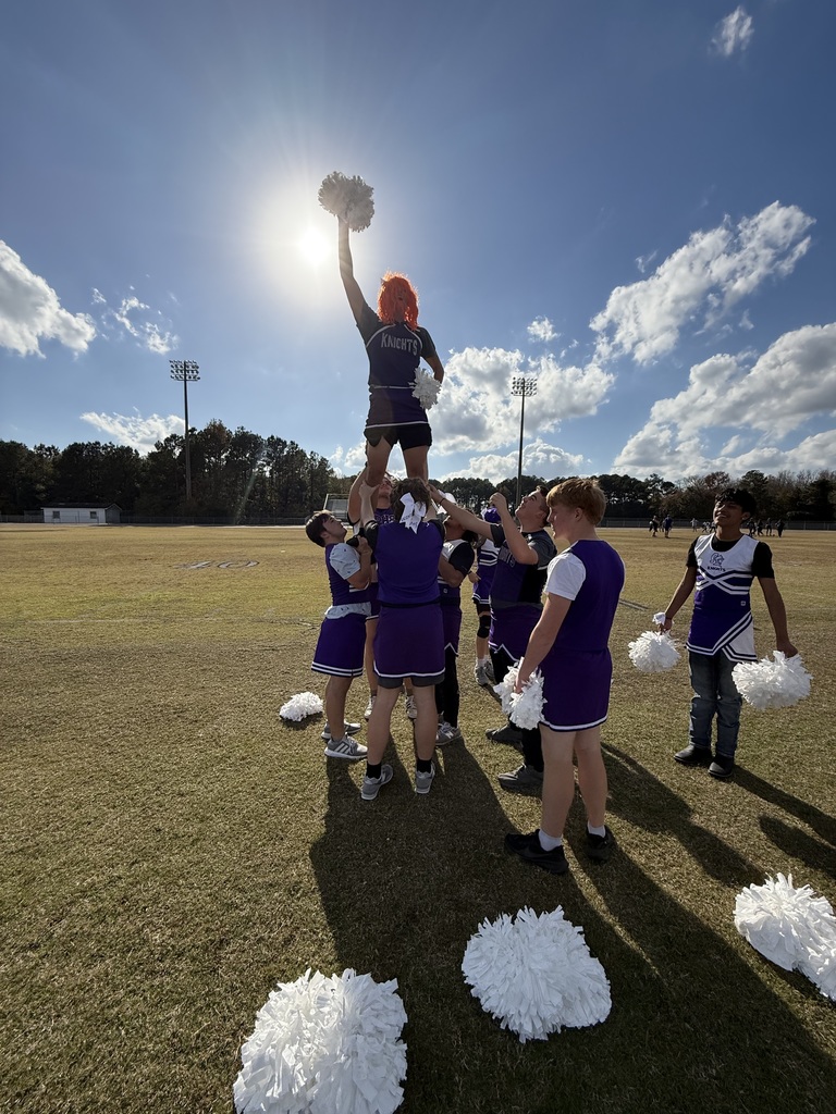 Cheerleaders perform a stunt pyramid outdoors; the top flier raises a pom-pom toward the bright sun.