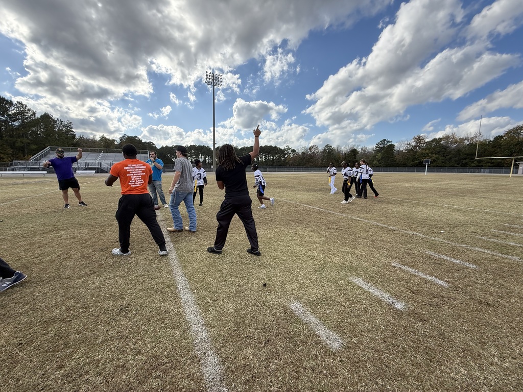 Coaches and flag football players on a field with one person pointing a finger to the sky.