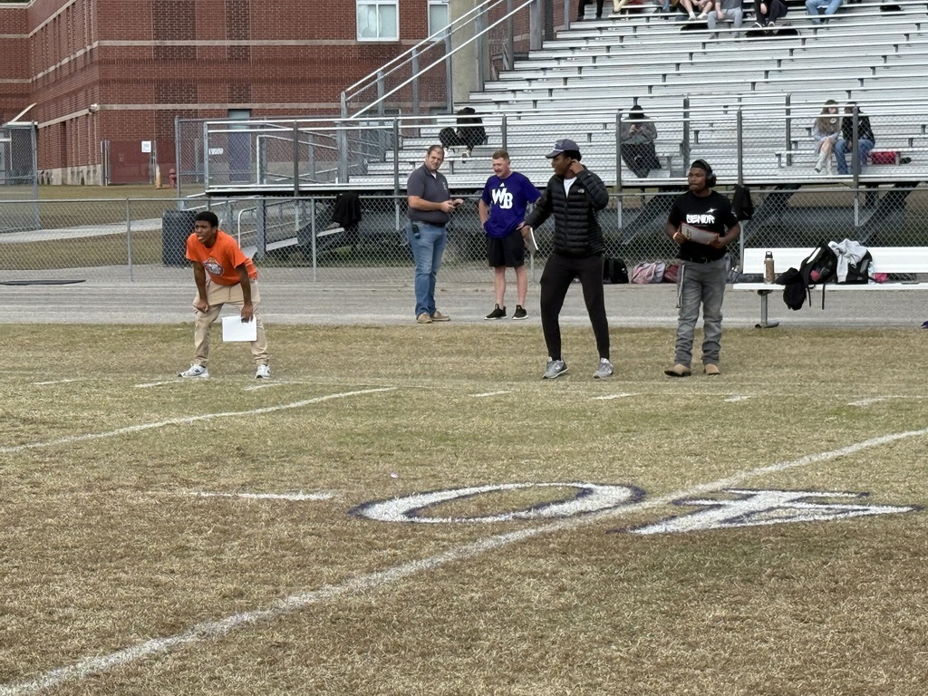 A flag football player in an orange shirt crouches in position on a grassy field with coaches watching nearby, set against a backdrop of stadium bleachers and a school building.
