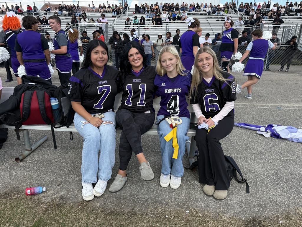 Four smiling girls in "West" or "Knights" jerseys sit on a bench in front of crowded bleachers