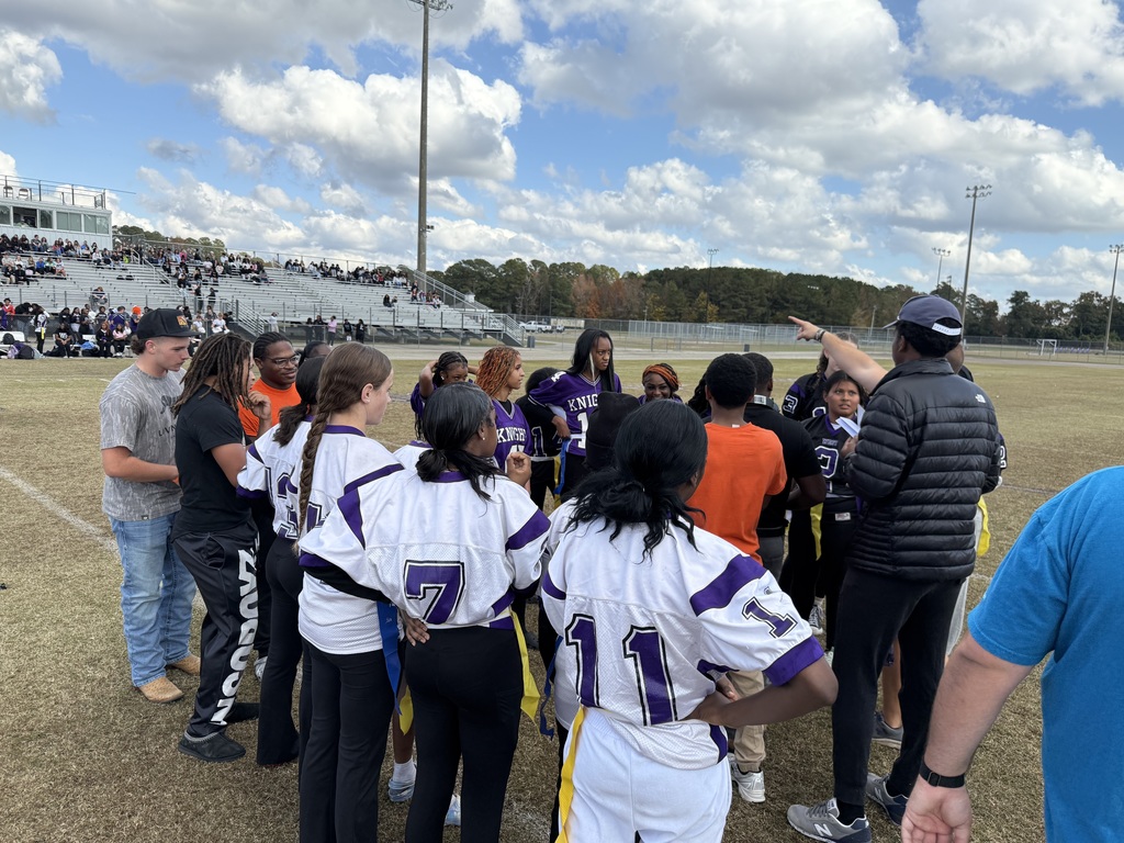 A flag football team in purple and white jerseys huddles with coaches on a field with spectators in the background.