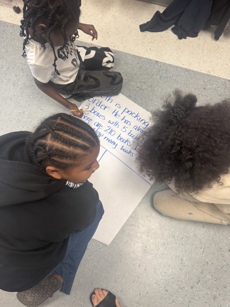Three students sit together on the floor working on a chart paper word problem. A student writes in pencil while the others watch and discuss what to do next. Classroom items and the teacher’s feet are visible nearby.