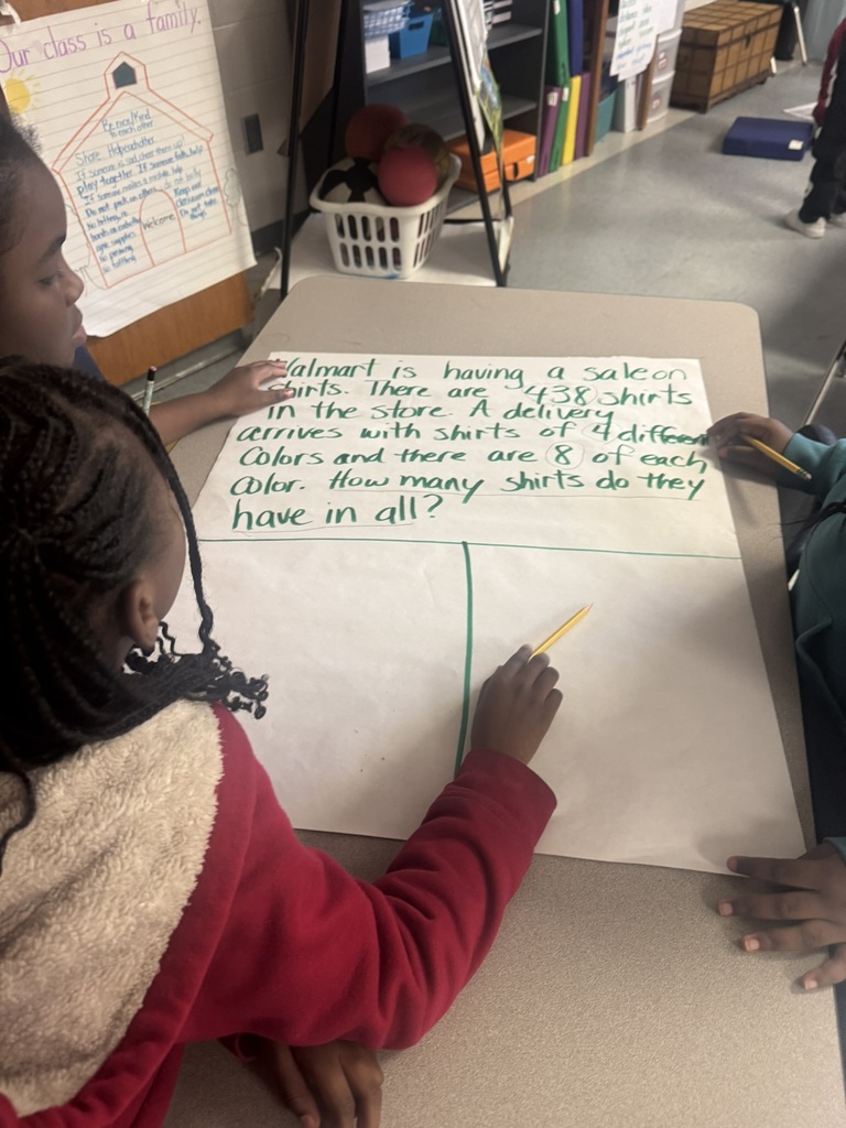 Two students sit and stand around a table, solving a multi-step word problem written on chart paper. One child points at the problem while another holds a pencil over the empty workspace. Classroom posters and supplies sit in the background.