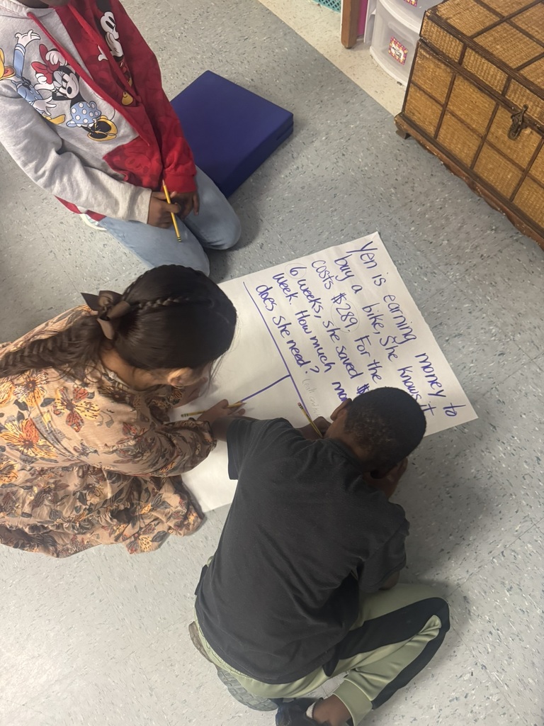 Three elementary students sit and kneel on the classroom floor working together on a large sheet of chart paper with a two-step word problem written in blue marker. One child writes while the others watch closely and discuss. Their workspace is surrounded by classroom materials and storage bins.