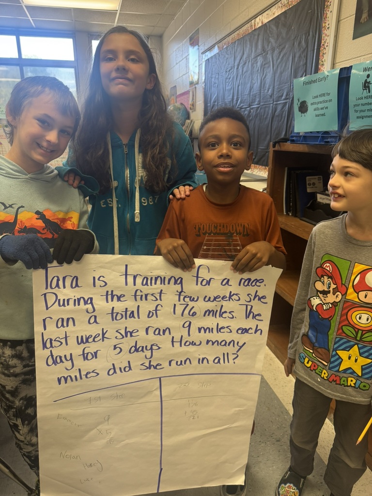 Small groups of smiling students hold up their completed chart paper showing their two-step word problems and written solutions. Each group stands proudly in their classroom, displaying their math work and teamwork.