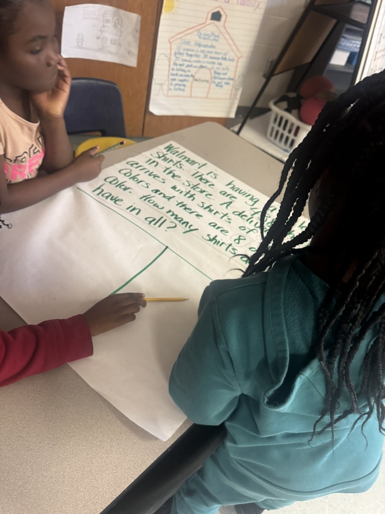 wo students sit and stand around a table, solving a multi-step word problem written on chart paper. One child points at the problem while another holds a pencil over the empty workspace. Classroom posters and supplies sit in the background.