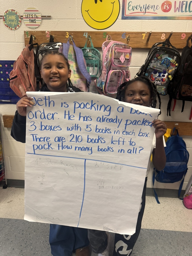 Small groups of smiling students hold up their completed chart paper showing their two-step word problems and written solutions. Each group stands proudly in their classroom, displaying their math work and teamwork.