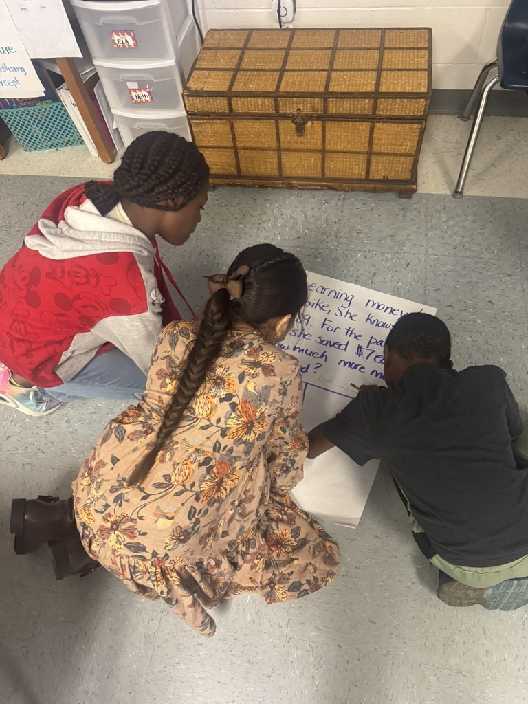 Three elementary students sit and kneel on the classroom floor working together on a large sheet of chart paper with a two-step word problem written in blue marker. One child writes while the others watch closely and discuss. Their workspace is surrounded by classroom materials and storage bins.