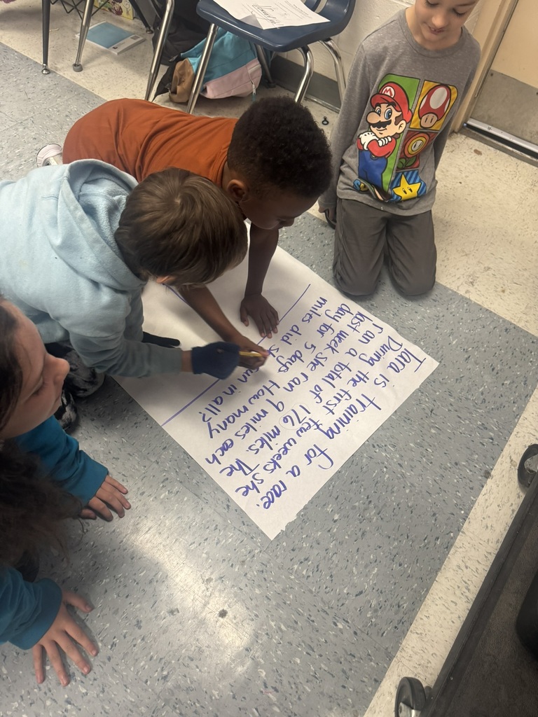 A small group of students leans over a large piece of chart paper on the floor, reading and solving a word problem written in blue marker. One student writes while the others point and follow along. Classroom chairs and bags are visible around them.