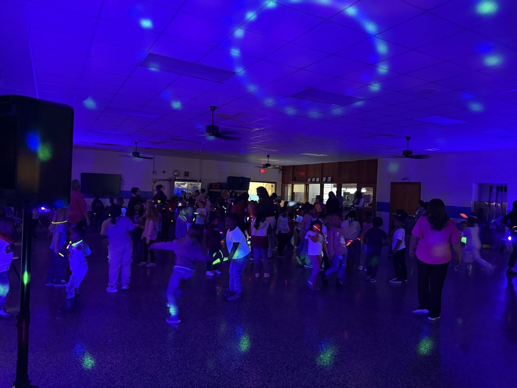 A large group of students dances in a darkened room illuminated with blue and green glow lights. Many children wear glow necklaces and bracelets as they move around the space.