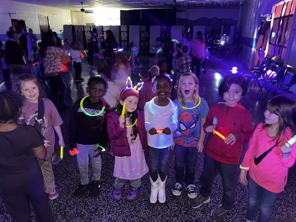 A group of eight children stands in a row, smiling and holding glow sticks while glow lights illuminate the room. Other students dance and move around in the background.