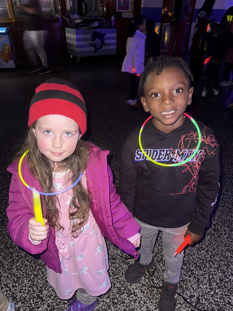 Two young children stand close together in a dimly lit room decorated with glow lights. Both wear glow necklaces and hold glow sticks, smiling at the camera. Other children are dancing in the background.