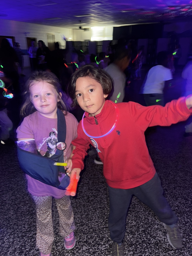 Two children stand side by side under black lights, wearing glow necklaces and holding glow sticks. One child has their arm in a sling, and both smile as other students dance behind them.