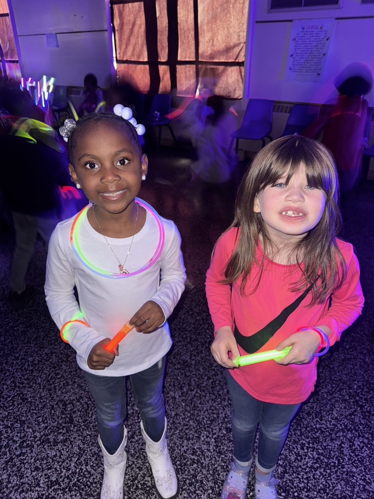 Two children stand together smiling in a glow-lit room. Both wear bright glow necklaces and hold glow sticks. Other students dance in the colorful lighting behind them.