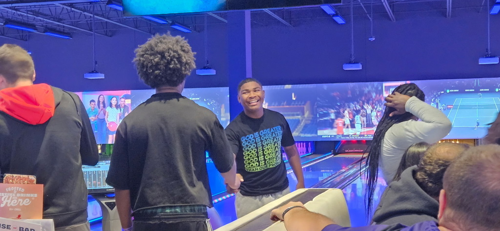 A young man smiles and shakes hands with a friend in front of bowling lanes and large video screens in a bowling alley