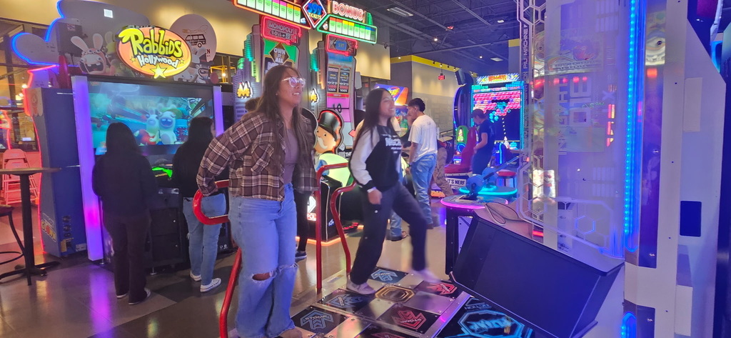 Two teenage girls smile while playing a dance-style rhythm game in a busy, colorful arcade.